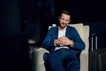 Handsome entrepreneur businessman, business traveler in business casual suit with a luggage at business trip, rests on an armchair in a lounge area of a luxury hotel, smiles looking at camera