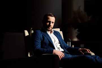 Fototapeta na wymiar Attractive self-confident business traveler, handsome successful businessman, entrepreneur resting in armchair in hotel lobby and confidently looking at camera