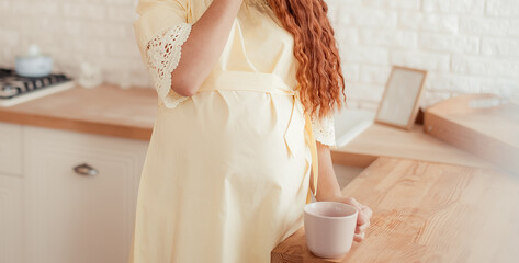 Fototapeta na wymiar A young pregnant girl in a delicate dress stands in the morning in the kitchen