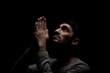 Headshot of serious confident young man praying in dark room. Lit from above and looking up