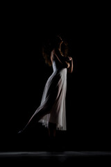 Girl with curly hair making ballet poses. Side lit silhouette of ballerina in white dress and black boots against black background.