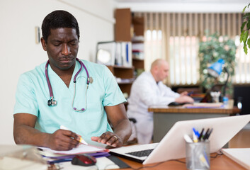 Experienced african american physician filling up medical forms on laptop while sitting at table in...
