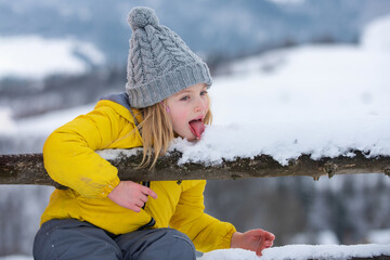 Winter girl eating snow outdoor. Funny christmas kids holiday. Kids cold and flu concept. Kid lick snow with tongue. Children wearing warm winter clothing in snow weather. - obrazy, fototapety, plakaty
