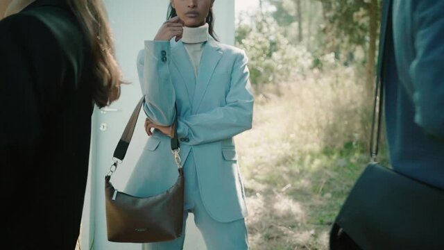 Two models moves away discovering a beautiful black woman pose for the camera wearing trendy suit and bag. Behind her there is a white single door standing