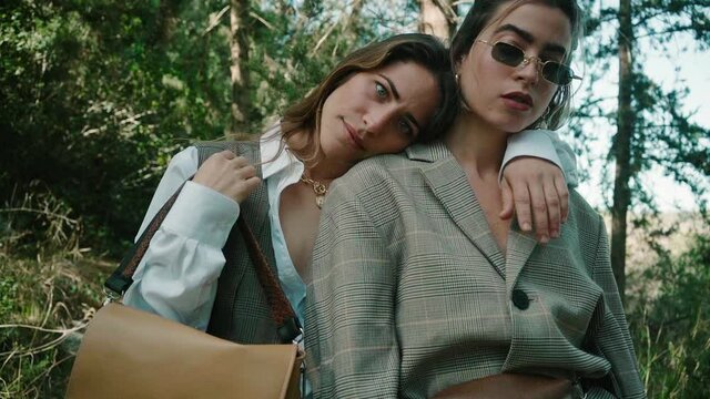 two beautiful women models posing for camera wearing trendy clothes, sun glasses and bags in a forest. One of the models hugging the other