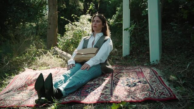 A beautiful woman sits on a carpet in the forest wearing trendy clothes and a hand bag
