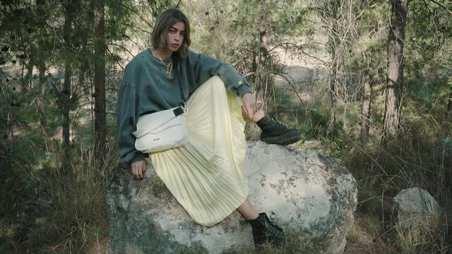 A beautiful woman pose for the camera sitting on giant rock wearing trendy outfit and hand bag in a forest