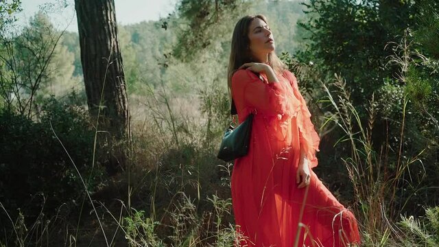 A beautiful girl in a dreamy peach-colored dress holds a handbag and poses for the camera in a forest