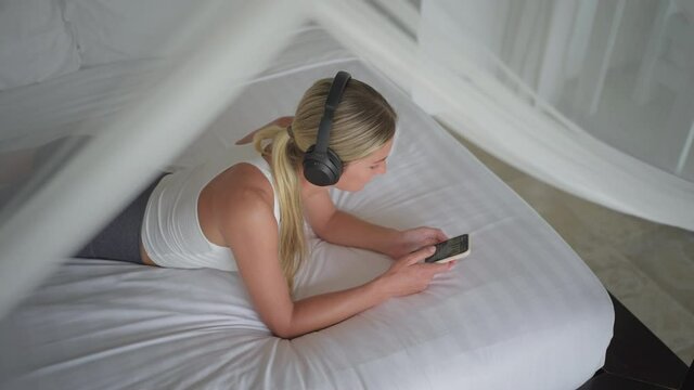 Woman listening music on wireless headphones in bed while scrolling on smartphone