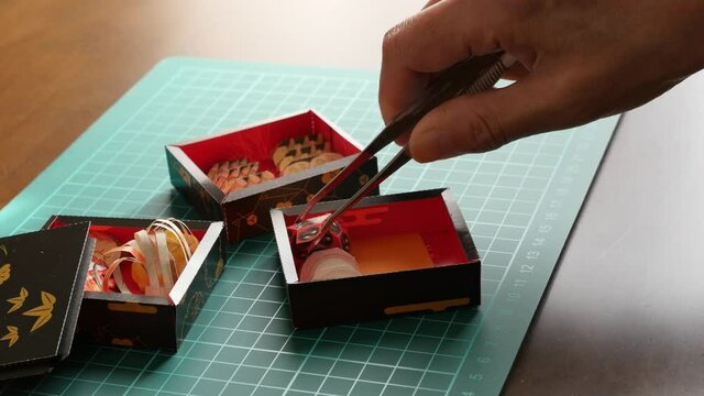 ペーパークラフトのおせち料理をピンセットでつまんで重箱に盛り付けてゆく
