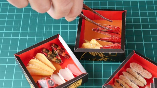 ペーパークラフトのおせち料理をピンセットでつまんで重箱に盛り付けてゆく
