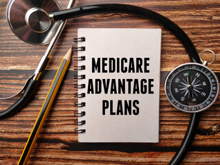 Notebook written with text MEDICARE ADVANTAGE PLANS with stethoscope, pencil and compass on a wooden background. - obrazy, fototapety, plakaty
