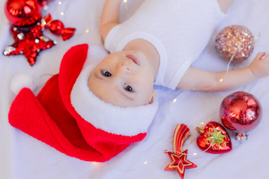 baby in a white bodysuit and a Santa hat is lying on his back on a white sheet surrounded by red Christmas tree toys. winter, new year. space for text. High quality photo