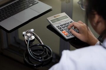 Female doctor with stethoscope using calculator and work on laptop computer at office desk in medical clinic or hospital. Medical cost and fee concept.