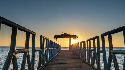 Obraz premium Sunrise over the Red Sea. A wooden path with a metal railing goes over the water to the canopy. The rays of the sun illuminate the sky in orange. Egypt