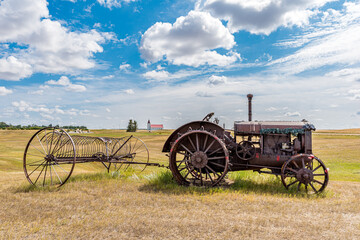Fototapeta na wymiar Antique tractor with steel wheels and lugs pulling a hay rake with St. Anthony Roman Catholic Church in Illerbrun, SK in the background