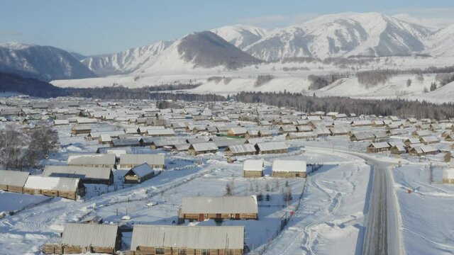 Aerial drone shot over Hemu Village, Xinjiang, China on a bright winter day. Snow covered wood cabins and beautiful mountains.