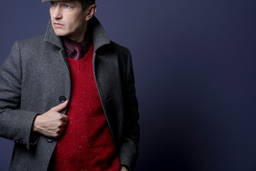 A man in a knitted red waistcoat, a gray coat and a tie a blue background.