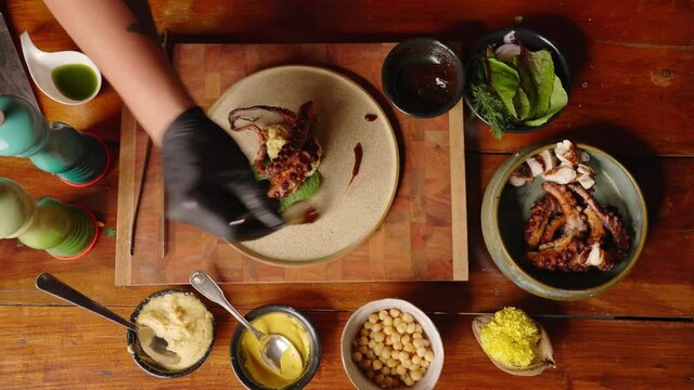 Top down view of a sophisticated professional fine dinning chef plating the smoked octopus and drizzle extra virgin olive oil to complete the dish.