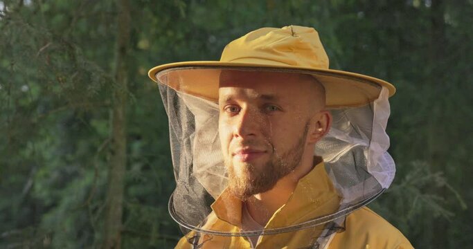 Smiling male beekeeper in the forest in the morning with the rising sun, wearing a yellow overalls face net to protect against insect bites, the guy takes care of the bees