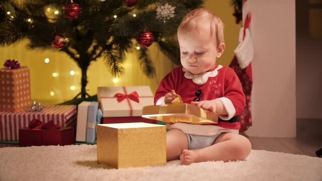 Cute baby boy unwrapping and looking inside Christmas present box under decorated Christmas tree. Families and children celebrating winter holidays.