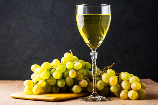 a glass of white wine and a brush of white grapes on a cutting board in the background on a black background