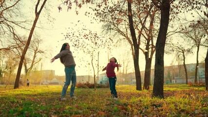 little child with her mother throw yellow leaves up in the autumn park at sunset, happy family, kid with mom make autumn golden leaf fall, enjoy fun fantasy of game, have fun in city rays of sunshine