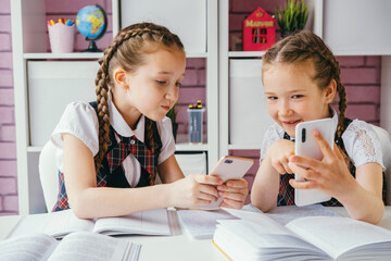 Two young female classmates are sitting at a desk and using smartphones. Modern technology concept. - obrazy, fototapety, plakaty