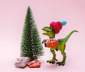 Funny plastic toy dinosaur wearing knitted hat with  present boxes and evergreen tree on a pink background.