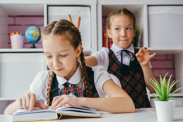 Lifestyle school portrait. Schoolgirl doing task sitting at desk, another girl doing bunny ears for her. - obrazy, fototapety, plakaty