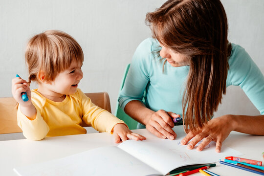 Mom teaches her little daughter to draw. Mother and daughter are sitting at a table and drawing.