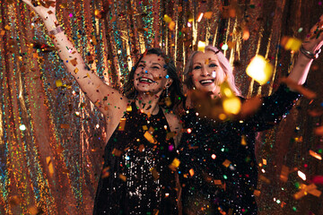 Two happy senior women in black dresses having fun under colorful confetti standing against a golden backdrop