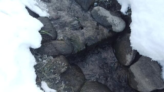 Steadicam shot of spring water flowing on the sand and bricks with snow, rocks and some dry grass around