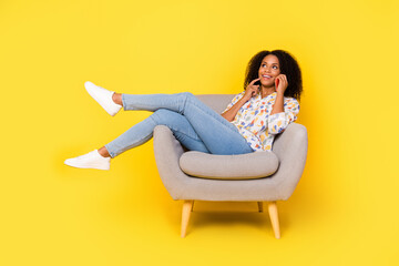 Full size photo of cute young wavy hairdo lady sit on armchair talk telephone wear shirt jeans footwear isolated on yellow background