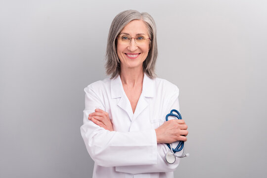 Photo of pretty cheerful senior woman doc dressed white uniform glasses smiling holding phonendoscope isolated grey color background
