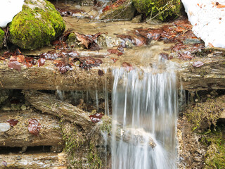 Small artificial waterfall. Water of a mountain stream flowing over wooden sill. Seen in the...