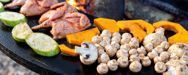 Close-up detail view of many tasty raw marinated quails and vegetables and mushrooms grilled cooked at round steel iron firepit hearth table surface brazier with burning firewoods. Barbecue yard home