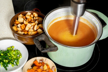 Hand blender in pot mixes soup, vegetable and fried piece of bread on board.