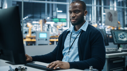 Car Factory Office: Portrait of Confident Black Male Chief Engineer Working on Desktop Computer. Professional Technician in Automated Robot Arm Assembly Line Manufacturing High-Tech Electric Vehicles