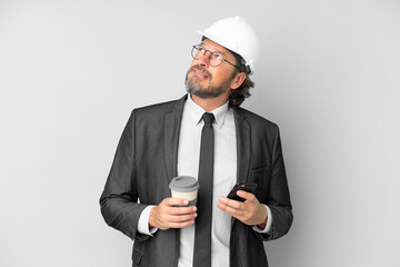 Young architect man with helmet over isolated background holding coffee to take away and a mobile while thinking something