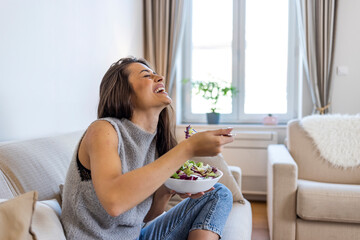 Photo of young woman enjoying a delicious salad while standing in her living room at home during the day. Young and happy woman eating healthy salad  with green fresh ingredients indoors