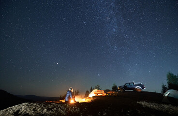 Lonely woman sitting on grass by burning fire near two tents and black SUV. Night camping on mountain hill under starfull sky. Tree tops and silhouettes of mountains afar on the background.