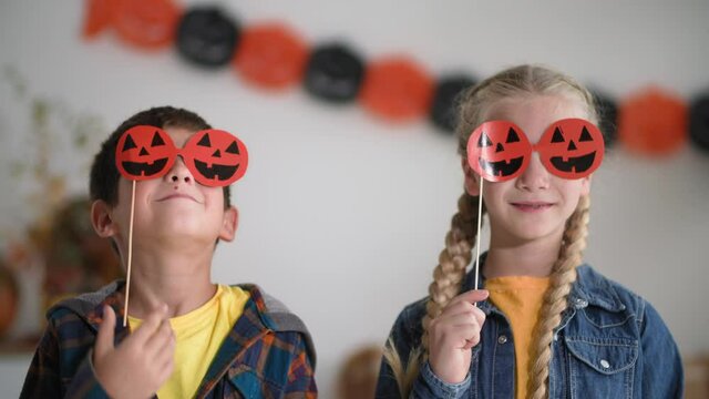 portrait of beautiful boy and girl putting on paper pumpkin glasses during halloween party, smiling and looking at camera and showing class
