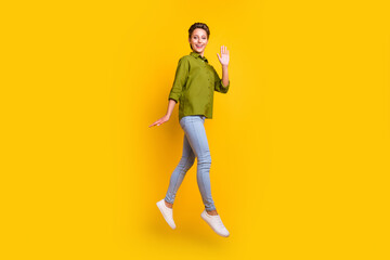 Fototapeta na wymiar Photo of friendly lady jump fly wave hand pose wear green shirt jeans shoes isolated yellow color background
