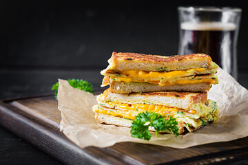 Roasted sandwich cheese and scrambled eggs on paper with coffee glass cup on wooden board. Dark background.