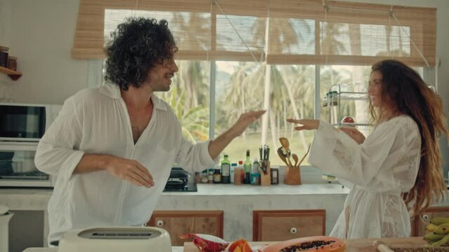 Young happy active married couple dancing laughing together cooking meals at home, carefree joyful husband and wife have fun cooking healthy breakfast food in boho styled kitchen