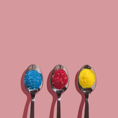 Three silver spoons arranged in line filled with colorful, shiny glitter piles – blue, red and yellow spangles on a pastel pink background. Creative minimal christmas flat lay concept. New Year idea