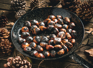 Roasted chestnuts in a chestnut pan (skillet with holes) on a table with autumn leaves and pine cones. Ready to eat. Traditional autumn food.