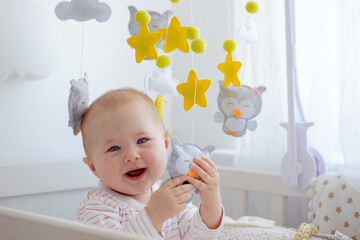 A laughing baby playing with toys of hanging mobile and looking at camera. Baby care, first skills, learning to sit.