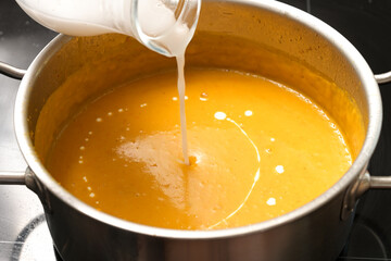 Pouring coconut milk into a vegetable cream soup from squash in a steel pot, vegan cooking in...